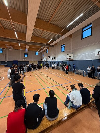 Jugendliche sitzen auf Bänken und stehen in einer langen Reihe in der Sporthalle