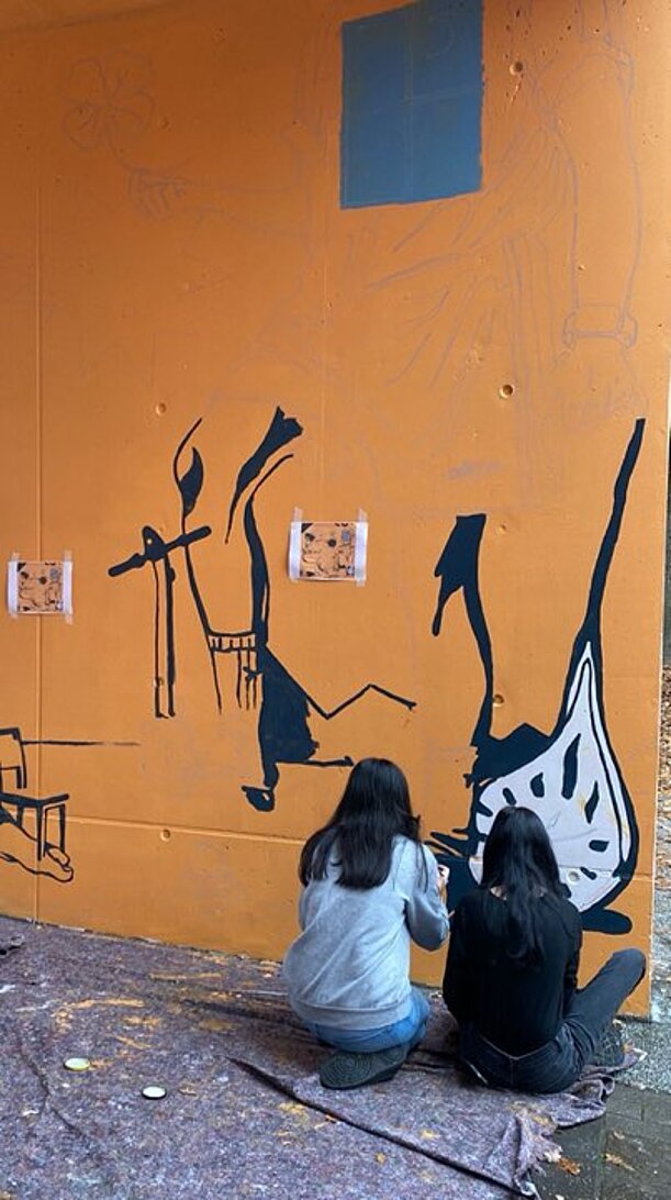 Zwei Jugendliche malen schwarze Umrisse auf eine orange Wand