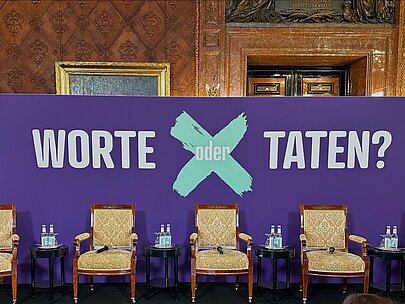Leere Podiumssessel vor dem Banner „Worte oder Taten?“ im Kaisersaal