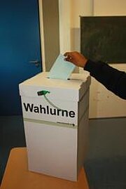 Hand wirft Stimmzettel in eine weiße Wahlurne bei der Juniorwahl