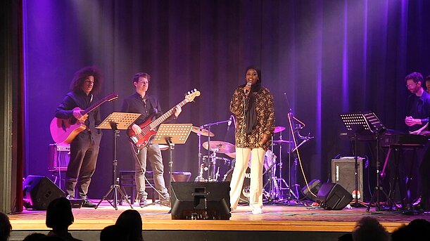 Sängerin in gemusterter Jacke vor Liveband auf violett beleuchteter Bühne