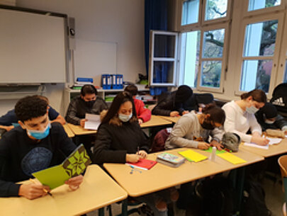 Schülerinnen und Schüler mit Masken gestalten im Klassenraum Briefe