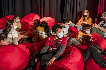 Grundschulkinder mit Masken lauschen auf roten Sitzsäcken dem Konzert