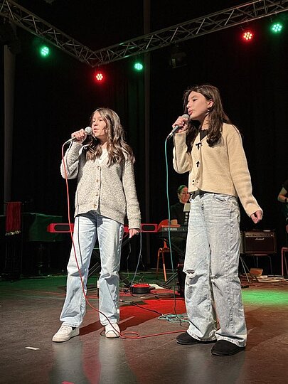 Zwei Schülerinnen in hellen Strickjacken singen im Duett beim Voice-Wettbewerb