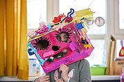 Kind trägt große pinke Maske aus Karton mit bunten Verzierungen