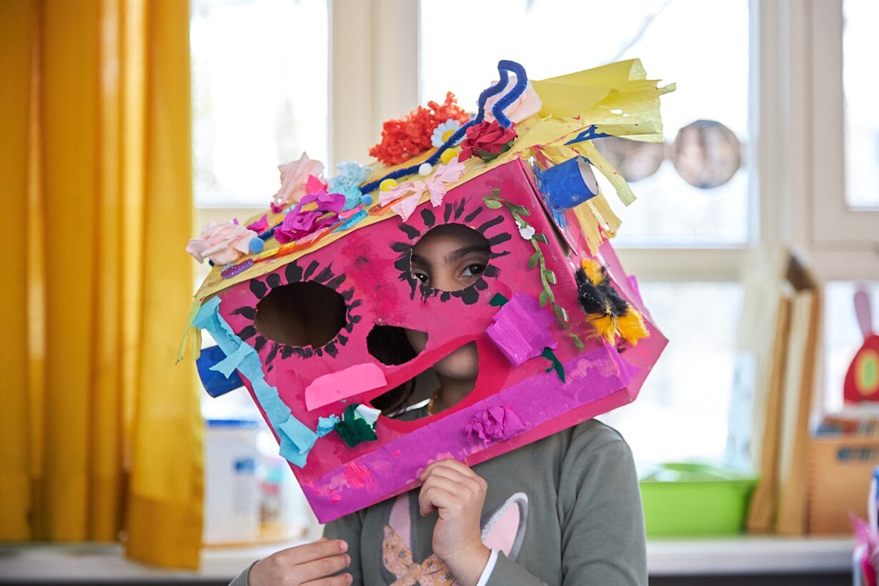 Kind trägt große pinke Maske aus Karton mit bunten Verzierungen