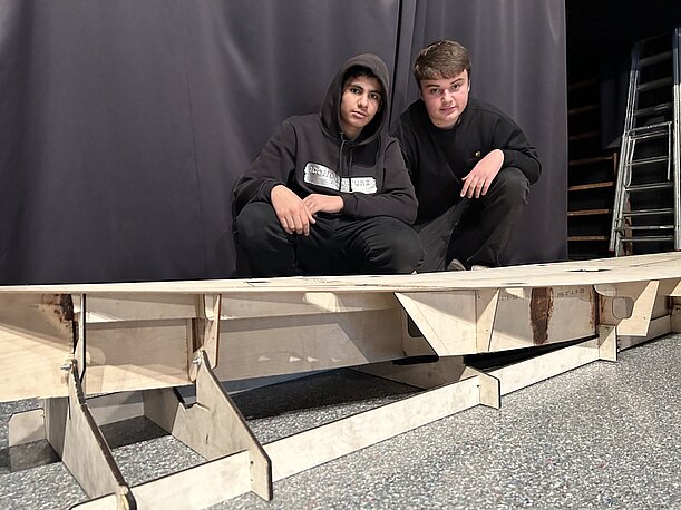 Zwei Jugendliche sitzen hinter einem selbstgebauten Holzmodell auf der Bühne