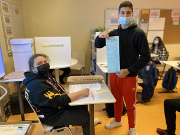 Schüler hält Stimmzettel der Juniorwahl hoch, daneben Mitschüler an der Wahlkabine