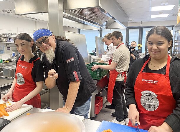 Schülerin schneidet Gemüse in der Großküche, daneben ein Koch mit Bandana