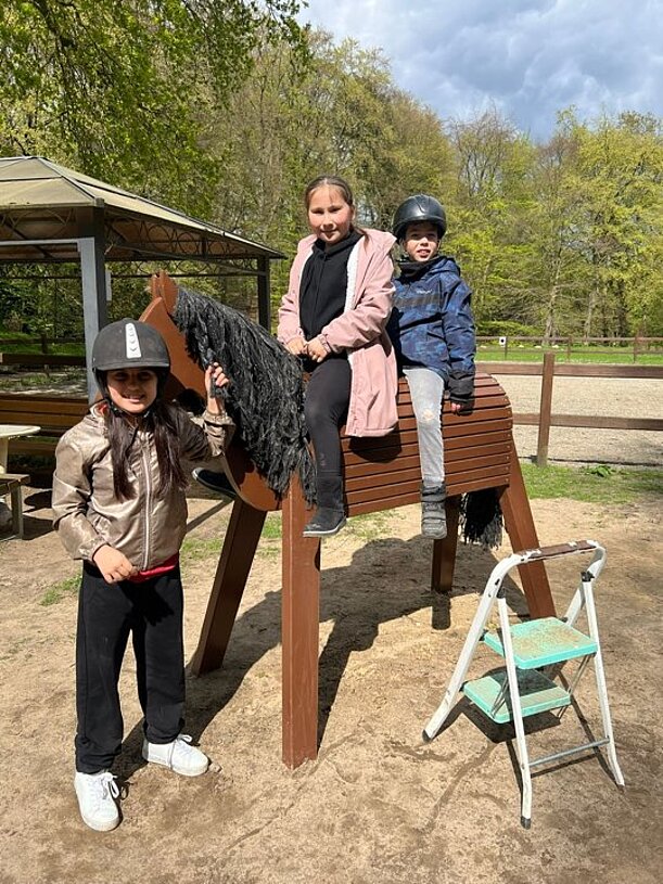 Kinder mit Reithelmen an einem Holzpferd auf einem Reitplatz
