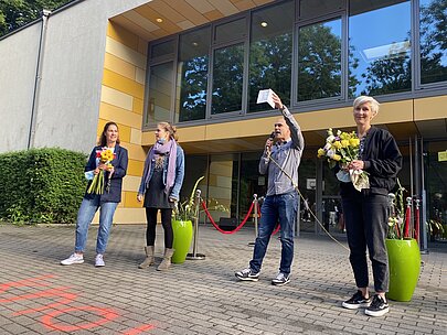 Lehrkräfte mit Blumen und Mikrofon bei der Begrüßung vor dem Schulgebäude