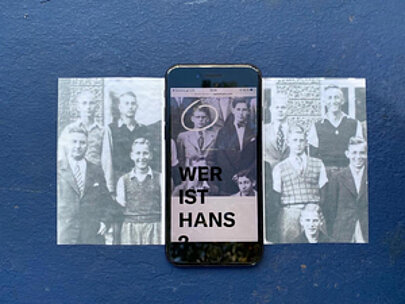 Smartphone mit Wer ist Hans zwischen historischen Gruppenfotos auf blauem Grund