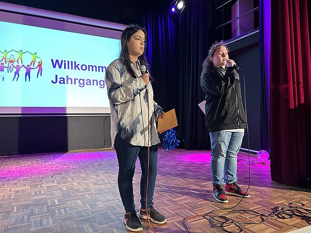 Zwei Schülerinnen moderieren mit Mikrofon vor Leinwand „Willkommen Jahrgang 9“