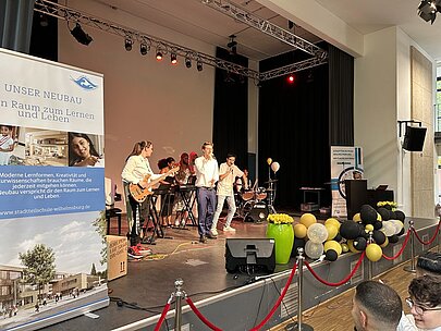 Band auf der Bühne neben einem Roll-up zum Schulneubau