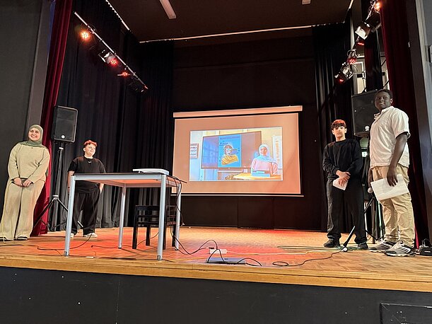 Präsentierende auf der Bühne neben Leinwand mit Videobild