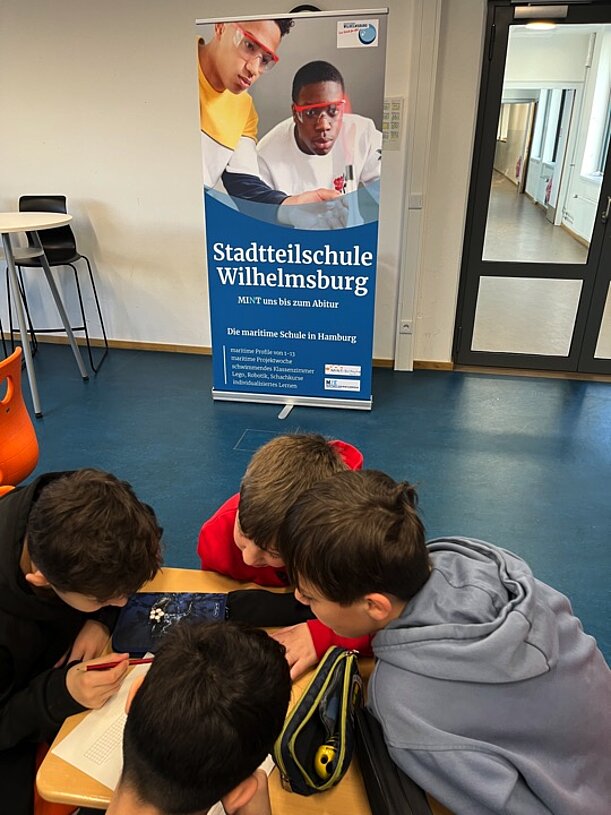 Kinder arbeiten gemeinsam an einem Tablet vor einem Schul-Roll-up