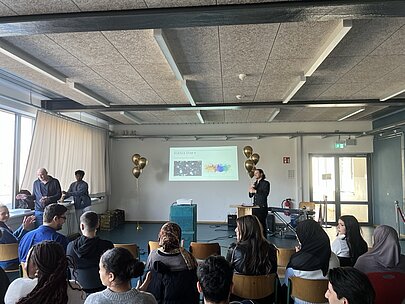 Saal mit Jugendlichen von hinten, vorne Leinwand und Moderator