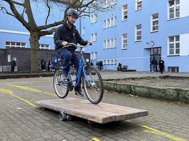 Schüler mit Helm fährt mit dem Fahrrad über eine Holzrampe auf dem Schulhof
