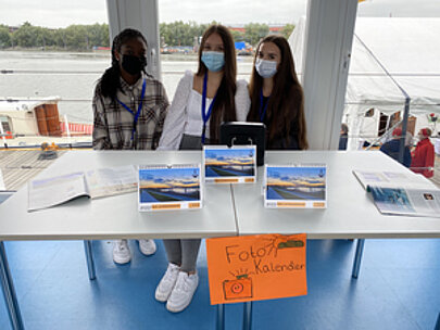 Drei Schülerinnen an einem Stand mit Fotokalendern auf dem Ponton