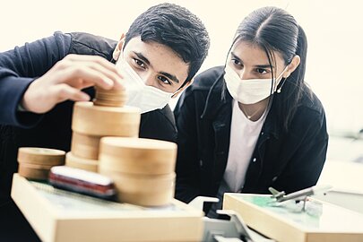 Schüler und Schülerin mit Masken stapeln Holzscheiben auf ein Modell