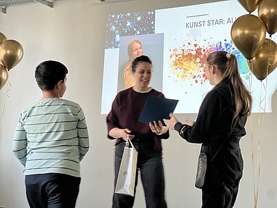 Lehrerin überreicht Urkunde vor Leinwand „Kunst Star“