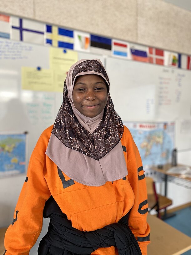 Schülerin in orangefarbener Jacke im Klassenraum mit Länderflaggen