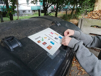 Hand klebt einen Aufkleber zur Mülltrennung auf eine Mülltonne