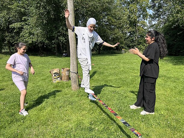 Schülerin balanciert auf einer Slackline zwischen Bäumen, begleitet von Mitschülerinnen