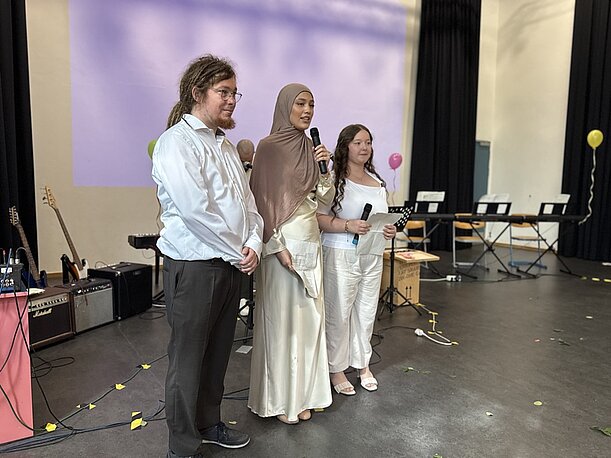Drei Moderierende mit Mikrofonen auf der Bühne der Aula