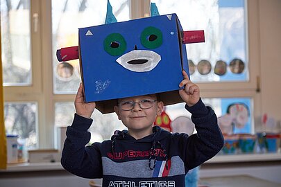 Junge hält blaue selbstgebastelte Kartonmaske über seinen Kopf