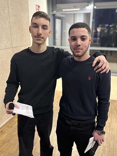 Zwei Moderatoren mit Mikrofon und Moderationskarten posieren im Foyer