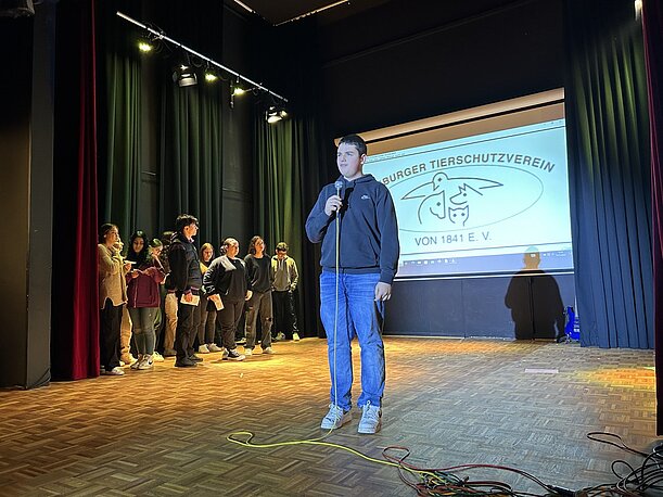 Schüler spricht vor Folie zum Tierschutzverein, Klasse wartet am Bühnenrand