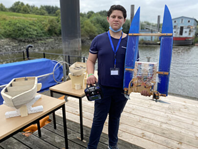 Schüler mit Fernsteuerung zeigt selbstgebautes blaues Modellboot am Spreehafen