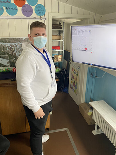 Schüler mit Maske steht in einem Raum neben Bildschirm mit Präsentation