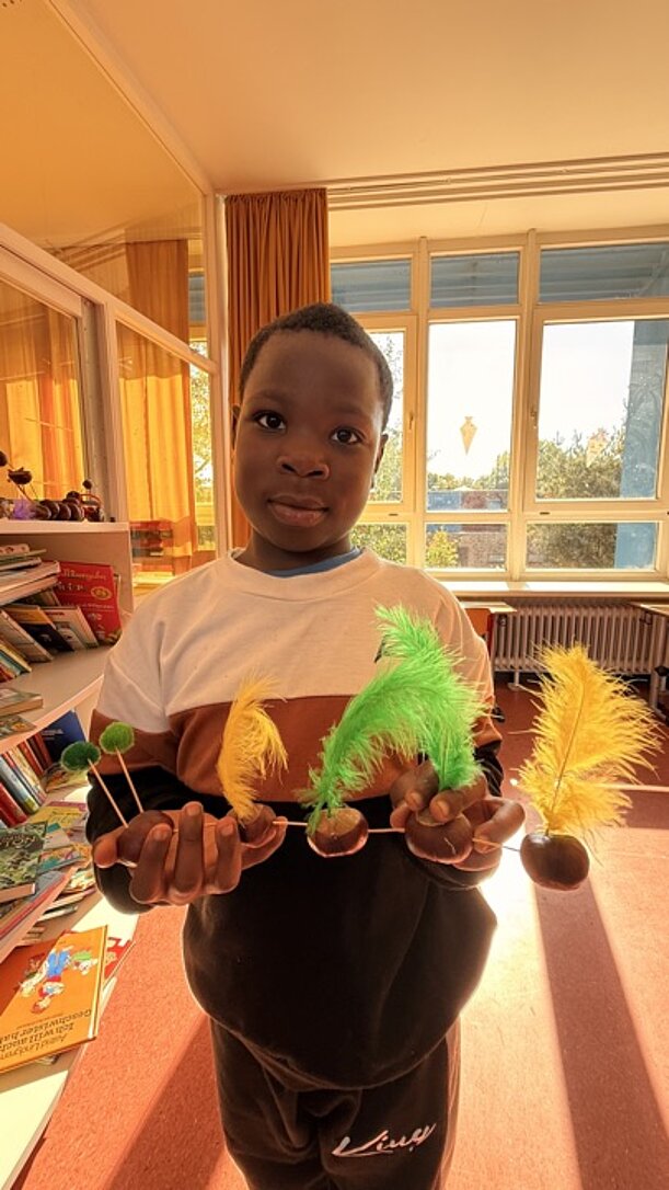 Junge zeigt Kastanienfiguren mit bunten Federn im Klassenraum