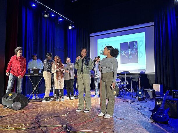 Jugendliche mit Mikrofon vor Leinwand und Bandinstrumenten bei der Profilpräsentation