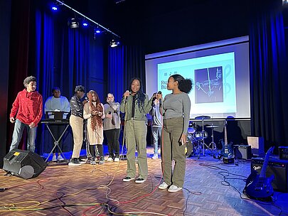 Jugendliche mit Mikrofon vor Leinwand und Bandinstrumenten bei der Profilpräsentation