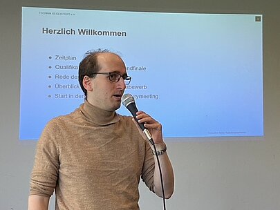 Tom Reinsdorf moderiert mit Mikrofon vor der Folie „Herzlich Willkommen“