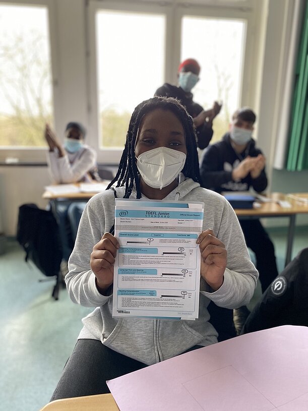 Schülerin mit Maske hält ihr Englisch-Zertifikat hoch