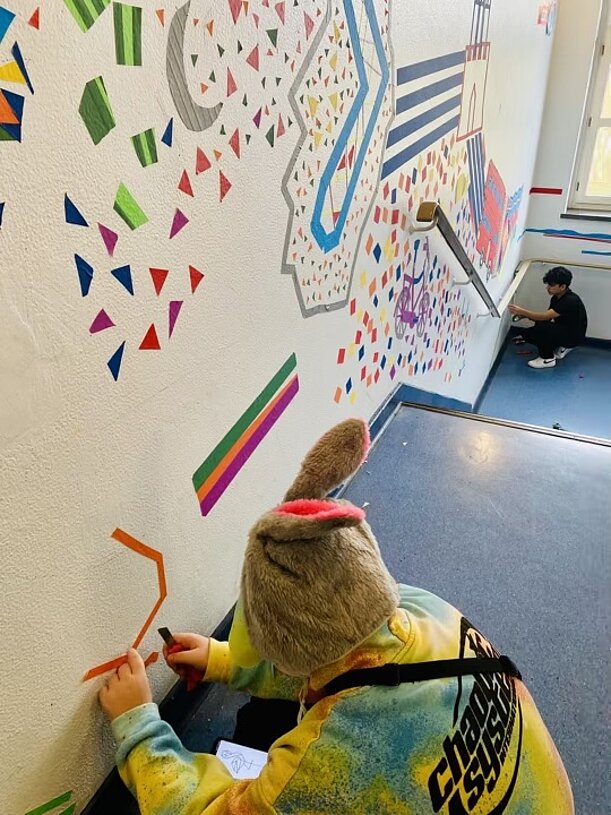 Schülerin klebt farbige Formen an eine große Tape-Art-Wand