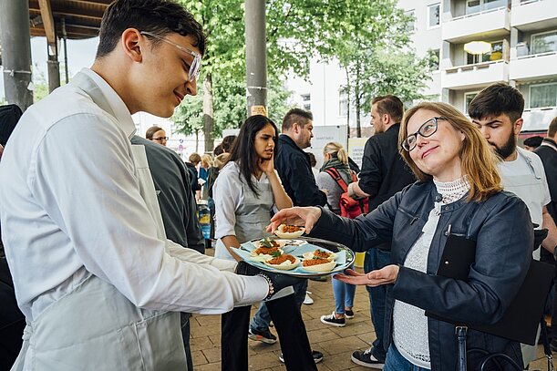 Schüler in Kochjacke reicht Besucherin einen Teller mit Essen