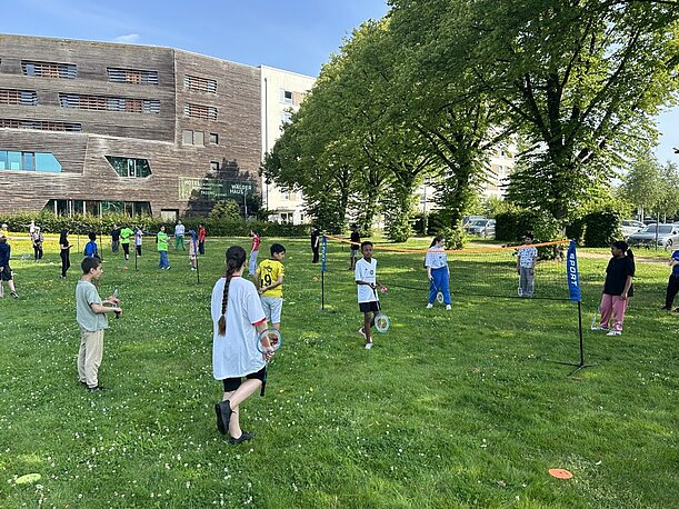 Kinder spielen Badminton auf einer Wiese vor einem Gebäude