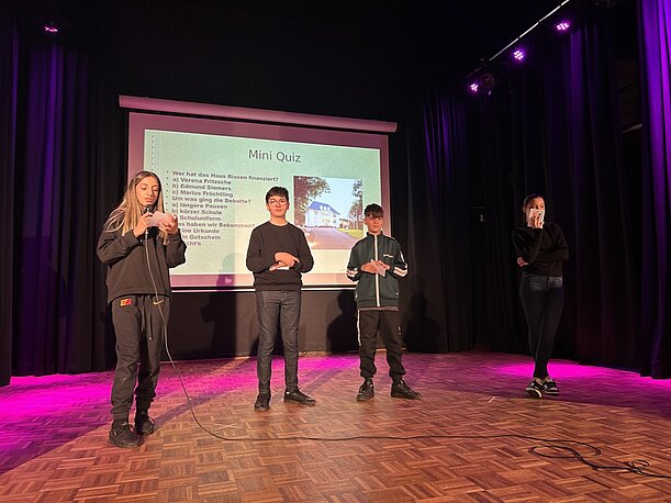 Schülerinnen und Schüler moderieren ein Mini-Quiz auf der Bühne der Aula