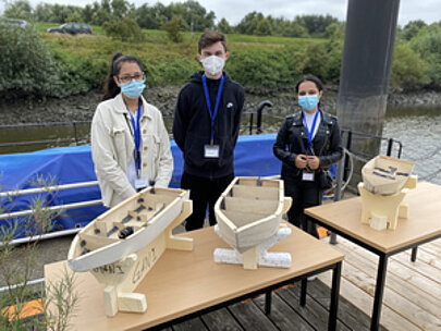 Drei Jugendliche mit Masken hinter Tischen mit Holzmodellbooten am Spreehafen