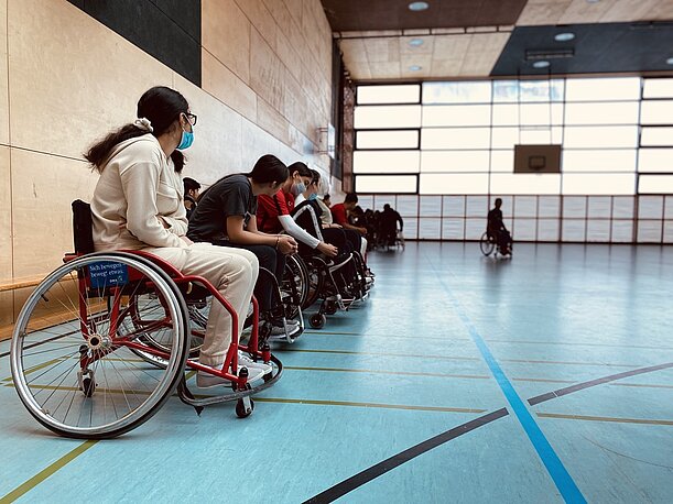 Schülerinnen und Schüler sitzen in Sportrollstühlen am Rand der Turnhalle