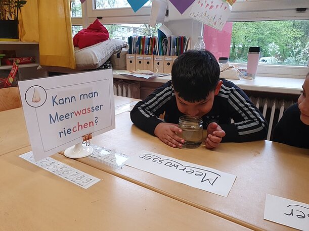 Remzi riecht an einem Glas Meerwasser neben dem Schild „Kann man Meerwasser riechen?“