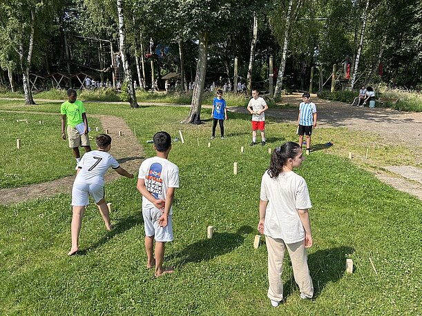 Kinder spielen Kubb auf einer Wiese unter Birken