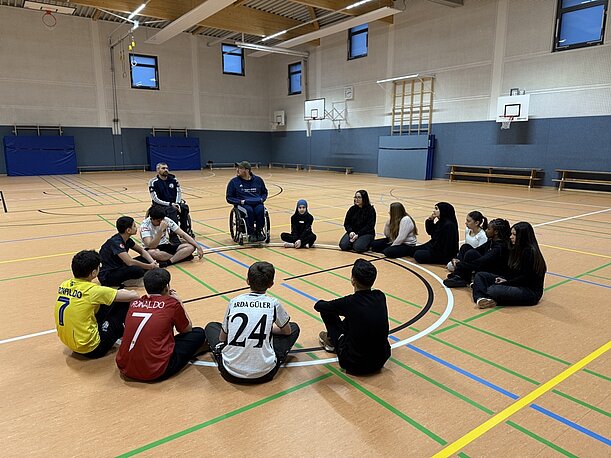 Jugendliche sitzen im Kreis mit Rollstuhlbasketballern zur Reflexion in der Halle