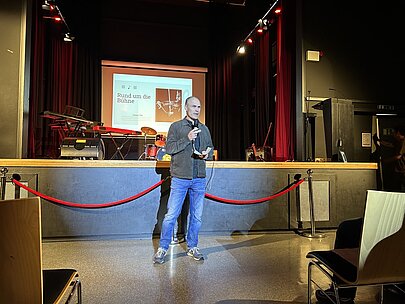 Lehrer begrüßt das Publikum in der Aula vor der Leinwand zur Profilvorstellung