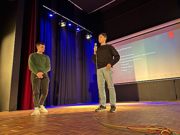 Zwei Schüler präsentieren mit Mikrofon vor Leinwand in der Aula